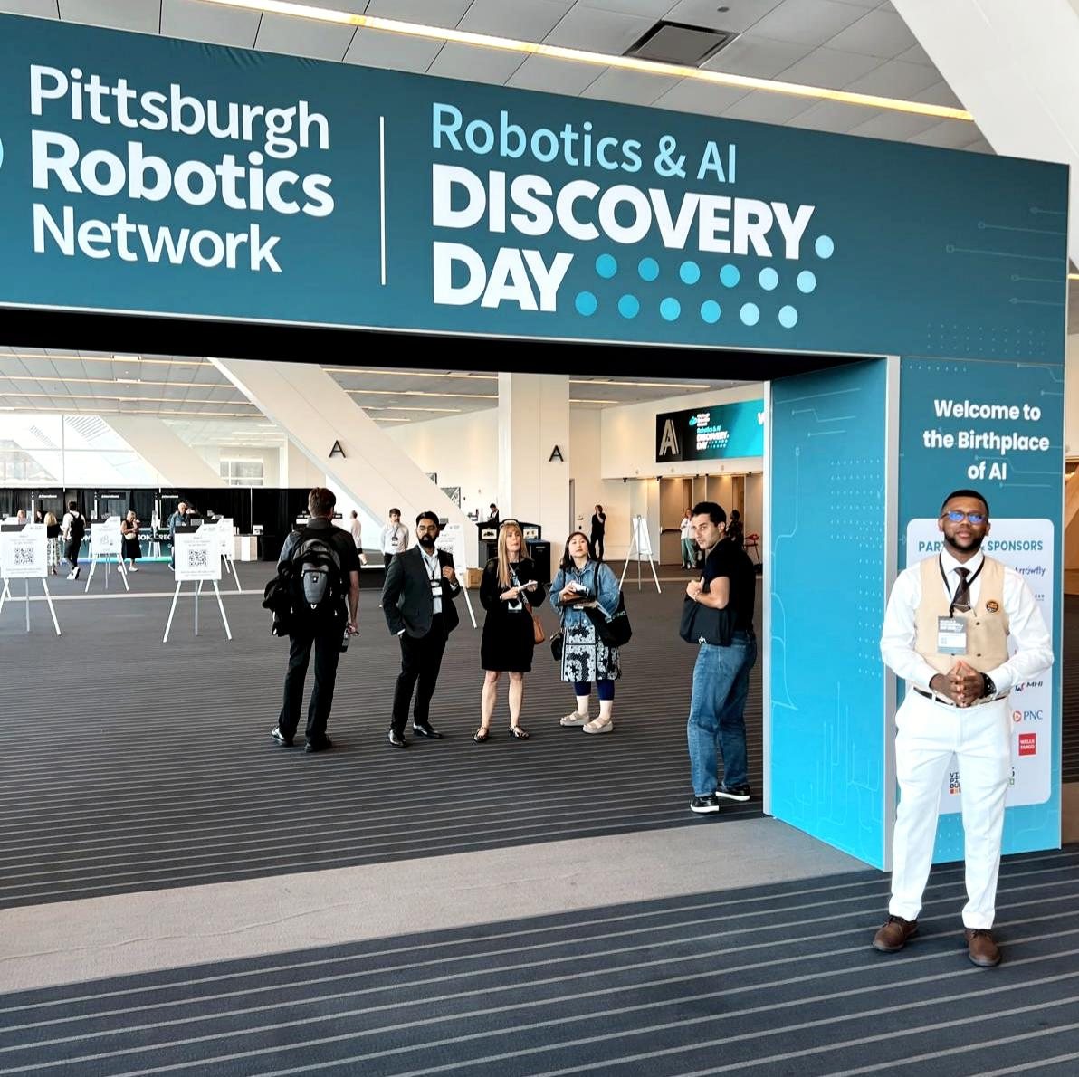 Johnson Oluwadele at the Pittsburgh Robotics Network's Robotics and AI Discovery Day entrance, beside a sign reading Welcome to the Birthplace of AI.